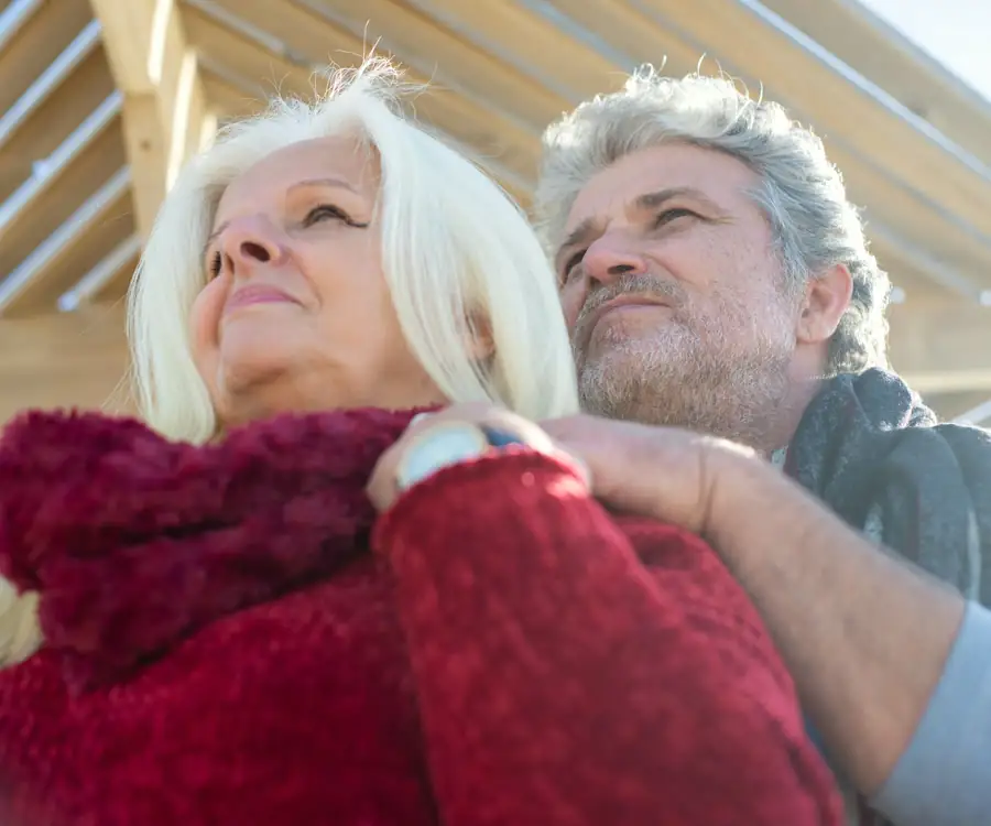 An older couple embracing under a wooden pergola in warm daylight, the woman with white hair looking up and smiling.
