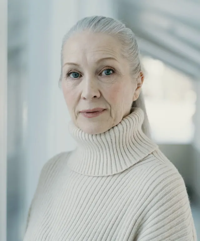 Portrait of Marguerite, a woman with gray hair pulled back, wearing a cream turtleneck against a soft light wall.