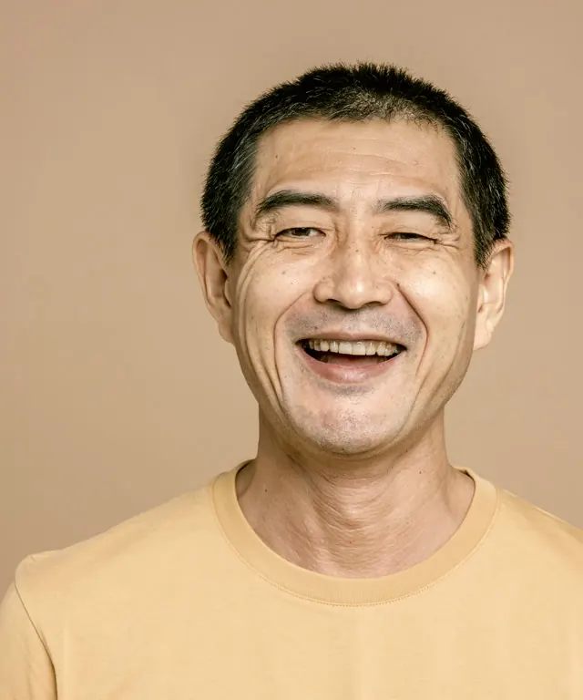 Portrait of Raymond, a smiling man with short dark hair in a mustard t-shirt against a warm tan backdrop.