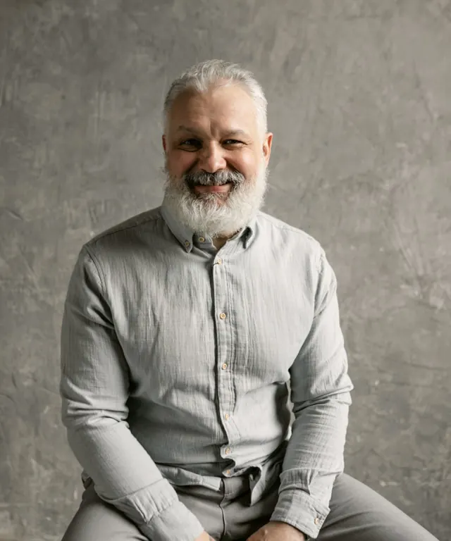 Portrait of Walt, a smiling older man with a gray beard in a light gray linen shirt against a neutral wall.